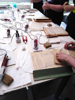 Das Foto zeigt einen Tisch, auf dem Materialien wie Zangen und andere Werkzeuge sowie Papiere mit mechanischen Sch&auml;den liegen. Den um den Tisch herumstehenden Studierenden wird gezeigt, wie sie Risse schlie&szlig;en k&ouml;nnen.