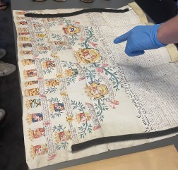 Das Foto zeigt einen kunstvoll gestalteten historischen Stammbaum einer Adelsfamilie, der der Einf&uuml;hrung in die Adelsarchivpflege diente. Eine behandschuhte Hand deutet mit ausgestrecktem Zeigefinger auf das Hauptwappen im Zentrum des Stammbaums.
