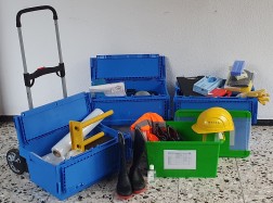 Auf dem Boden stehen drei blaue und eine grüne Kisten mit Werkzeug, Helm und Stiefeln.