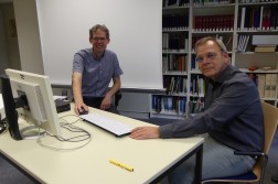 Foto: 2 Männer sitzen an einem Arbeitstisch mit PC vor Bücherregal