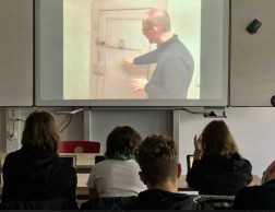 Im Vordergrund sind vier sitzende Schüler*innen, die auf eine Leinwand schauen. Dort sieht man die Videoaufnahme eines Mannes, der eine Zellentür erklärt.