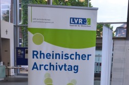 Bild von einem Aufsteller in einem Raum mit Glaswänden, auf dem Aufsteller ist die Aufschrift "Rheinischer Archivtag" zu lesen, darüber steht das Logo des LVR